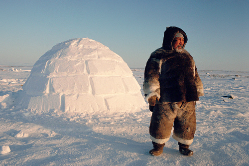 A man in a fur parka standing next to an igloo on snow